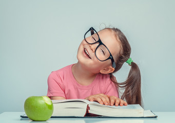 Wall Mural - cheerful beautiful cute little girl next to thick book and green apple looking at camera