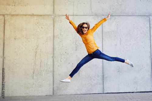 Photographie Joyful young lady jumping and raising arms in front of wall outside