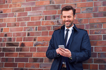 happy middle-aged businessman using phone and leaning on wall while outside