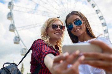 Portrait of two beautiful young girls taking selfie via smartphone camera standing against tall Ferris wheel in amusement park