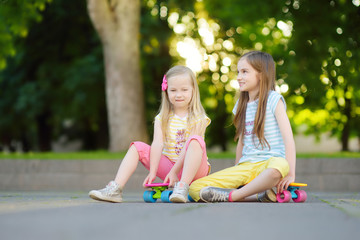 Two pretty little girls learning to skateboard on beautiful summer day in a park. Children enjoying skateboarding ride outdoors.