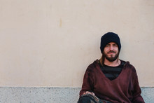 Portrait Of Young Homeless Man Sitting Against Blank Concrete Wall
