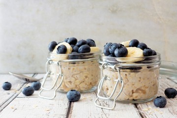 Wall Mural - overnight oats with fresh blueberries and bananas in jars on a rustic white wood background