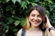 © shurkin_son - Headshot of cute charming teenage girl with and pretty set of features adjusting her hair and looking sideways with shy smile while standing outdoors at green wall and flirting with guy she likes