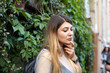 © shurkin_son - Outdoor picture of stunning fashionable hipster girl posing at green wall outdoors on sunny day, wearing wrist watch, nose-ring and backpack, looking down with thoughtful expression on her face