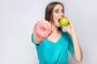 Beautiful young woman with freckles in green dress, eating green apple and holding pink donut. studio shot on light gray background