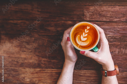 Plakat Filiżanka kawy latte w ręce Hipster Kobieta Lay na drewnianym stole w Cafe, widok z góry