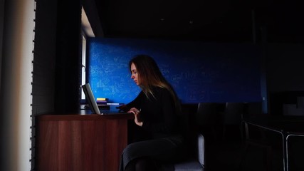 Wall Mural - Nice successful student writes essay and types text on computer keyboard and sits at table in modern library or cafe on winter day. beautiful European-looking woman with long blond hair dressed in