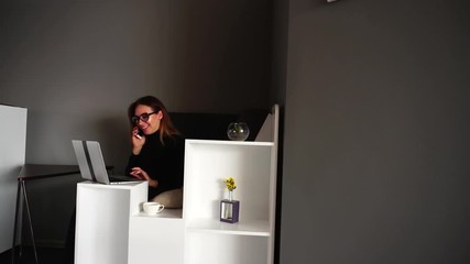 Wall Mural - Lovely female chatters on mobile with employee and discusses working moments, turns to laptop and works behind it, smiling and laughing, sitting at computer in stylish modern office during day
