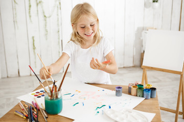 Having fun, joyful, smiling with teeth blonde seven-year-old girl dripping paint over white sheet of paper lying on a table. Creative child having fun, enjoying painting.