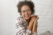 © wayhome.studio  - Pleased mixed race woman with curly hair wearing big eyewear and checkered shirt closing her eyes with pleasure while smiling broadly and keeping hands together, receiving good news from friend