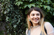 © shurkin_son - Positive cheerful teenage girl with long blonde hair wearing stylish clothes and accessories looking sideways with happy smile, hiding in shadow of green wall, having walk with fiends in public garden