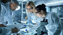 Team Of Medical Research Scientists Work On A New Generation Disease Cure. They Use Microscope, Test Tubes, Data Implementing Technology. Laboratory Looks Busy, Bright And Modern.