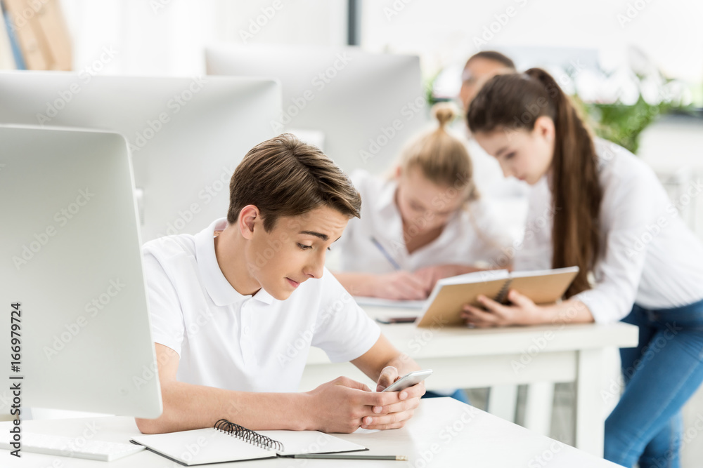 caucasian teenage boy using smartphone while sitting at table during lesson in classroom - obrazy, fototapety, plakaty 
