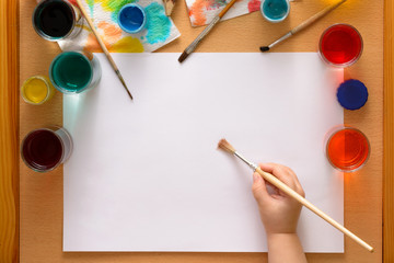 Preparing to drawing. Blank sheet among watercolors and brushes.Child's hand with brush above sheet