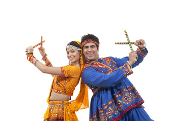 Portrait of happy young couple in traditional wear standing back to back while performing Dandiya Raas over white background