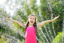 Adorable Little Girl Playing With A Sprinkler In A Backyard On Sunny Summer Day. Cute Child Having Fun With Water Outdoors.