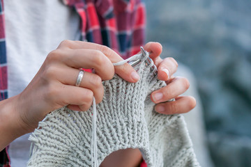 Close-up Young woman checkered red shirt and white t-shirt knits with knitting needles gray sweater from natural woolen threads, in the background stones