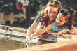 On a warm summer day, a mother, and her daughter joyfully set sail paper boats in a vibrant fountain, sharing precious moments and laughter by the water's edge.