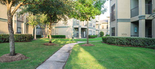 Wall Mural - Clean lawn and tidy oak trees along the walk path through the typical apartment complex building in suburban area at Humble, Texas, US. Grassy backyard, sunset warm light. Panorama.