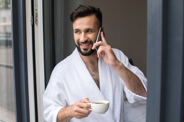 Wall Mural - handsome smiling bearded man in bathrobe talking on smartphone and drinking coffee at home