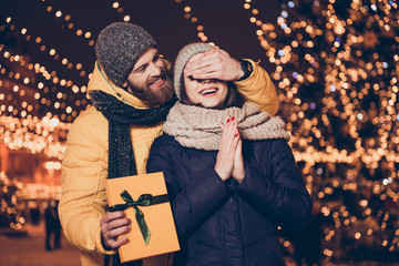 Have something for you dear! Smiling cute brunette is waiting for surprise from her lover, who closed her eyes with hand. Christmas eve outdoors, many lights around