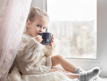 A Little Girl In The White Knitted Sweater Sits On A Window And Drinks From A Cup