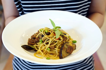 Hand holding a white plate of Green Curry Spaghetti with grilled purple eggplant (aubergine), roasted pleurotus eryngii mushroom and edamame decorated with basil, Vegan or vegetarian Food concept.