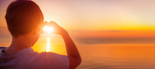 Happy little boy kid making heart with his hands over sunset sea background. Vacation concept. Summer holidays