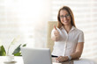 © fizkes - Attractive businesswoman looking at camera, showing thumbs up at workplace, pretty happy manager raising finger sitting at office desk with laptop, likes her job, acceptance approval sign, good idea
