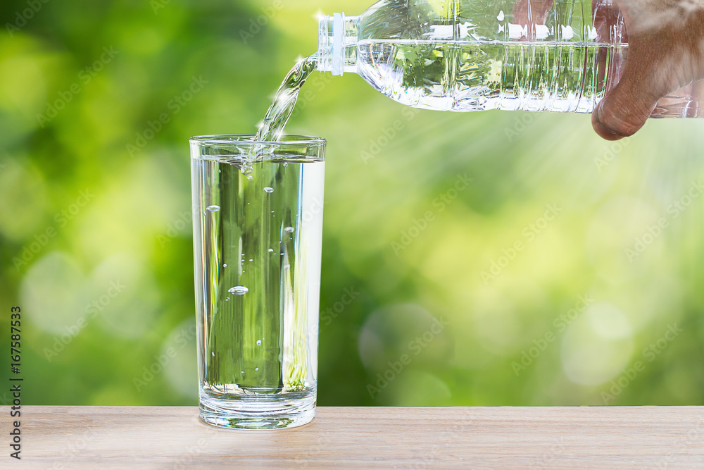 Man's hand holding drinking bottle water and pouring water into glass on wooden table on blurred green nature background with soft sunlight - obrazy, fototapety, plakaty 