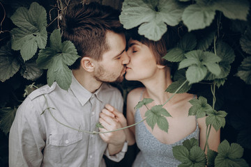 stylish hipster couple kissing in green leaves, holding hands. man and woman embracing, in love relaxing in summer park, picnic date. girl in fashionable modern dress. rustic wedding concept