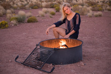 Blonde Young Woman Sitting By Fire Pit With Wind At Night With Flame In Desert