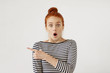 Amazed female model with red hair tied in knot, wearing stripe clothes, looking with bugged eyes and widely opened mouth, pointing with forefinger at copy space for your text or advertisment