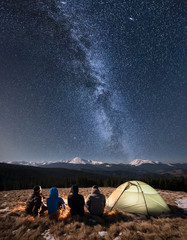 Wall Mural - Rear view of four people sitting together beside camp and tent under beautiful night sky full of stars and milky way. On the background snow-covered mountains. Long exposure