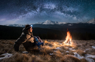 Wall Mural - Male tourist have a rest in the mountains at night. Guy with a headlamp sitting near campfire under beautiful night sky full of stars and milky way, and enjoying night scene