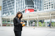 © Johnstocker - Business woman using phone with coffee in hand walking on the street with office buildings in the background