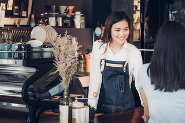 Wall Mural - Asian woman barista wear jean apron holding coffee cup served to customer at bar counter with smile emotion,Cafe restaurant service concept,Owner small business concept