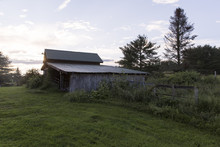 USA, Vermont. An Old Barn On The Turf Of A Farm With The Rising Sun Behind It.