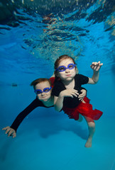 Two little girls swimming and posing underwater and watching me on the blue background. Portrait. Shooting under water. Vertical orientation