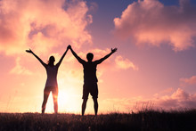 Sunset Silhouette Of Man And Woman With Arms In The Air Feeling Happy And Free. 