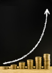 Money coin stack arranged as a graph on a wooden and write white chalk arrow indicates economic upturn on blackboard, concept of money growth