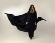 full length portrait of a blonde lady wearing black lace now and hooded cloak, standing pose isolated against creamy background.