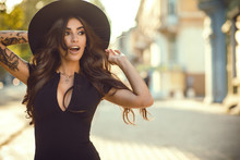 Portrait Of Gorgeous Glam Tattooed Lady With Long Wavy Hair And Beautiful Make Up Holding Her Trendy Fedora Hat For It Not To Be Blown Away By The Wind. Her Mouth Open With Surprise. Copy-space