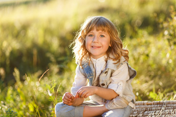 Wall Mural - A portrait of a cute little boy with blue eyes and long blond hair in a jeans west sitting on a basket outside at sunset an having fun