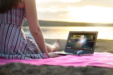 Young woman streaming a movie with laptop computer on beach at sunset. Watching film stream on imaginary online service outdoors. Video player on screen.