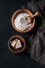 Fresh organic broken yeast cube, olive wood bowl of flour for baking homemade bread. With scoop, serving board, textile over black burned wooden background. Top view with copy space