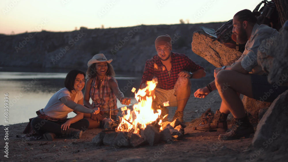 Group of multiracial people sitting around campfire grilling marshmallows and having fun on coast. - obrazy, fototapety, plakaty 