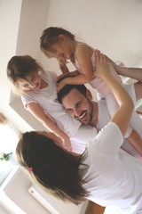 Wall Mural - Cheerful family playing together on bed. Parents spending free time with their daughters. Daughters sitting on fathers shoulders.