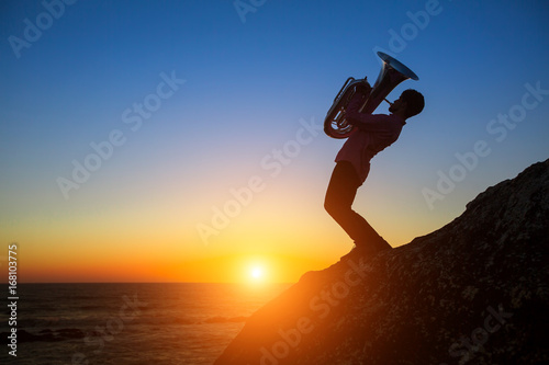 Plakat Sylwetka muzyk z Tuba na skalistym wybrzeżu morza podczas niesamowity zachód słońca.